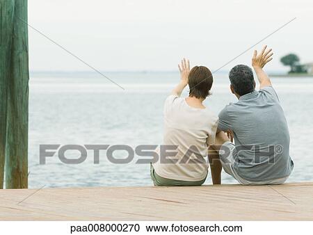 Stock Image - Couple sitting on edge of dock, waving. Fotosearch