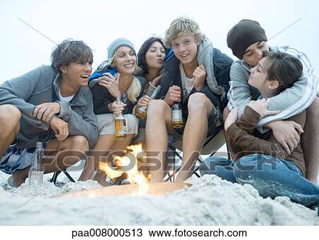 Stock Image - Group of young adults sitting around beach campfire, with bottles of beer. Fotosearch