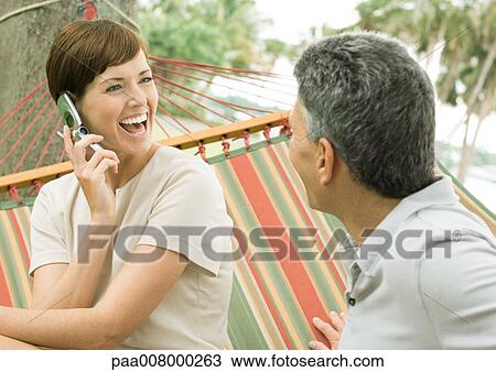 Stock Image - Mature couple lounging in hammock, woman using cell phone. Fotosearch