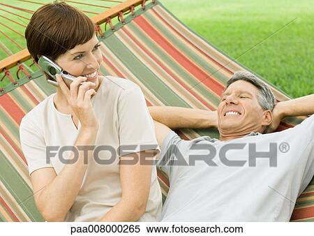 Stock Photography - Mature couple lounging in hammock, woman using cell phone. Fotosearch
