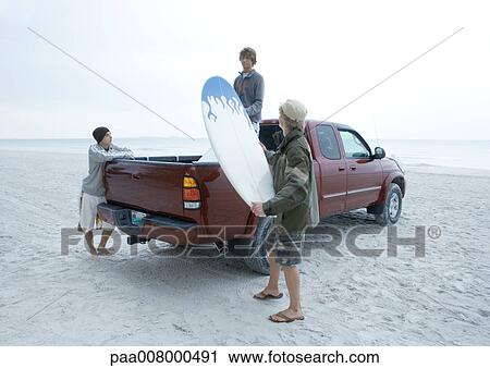 Stock Image - Three surfers taking surfboard out of back of pick-up truck, on beach. Fotosearch