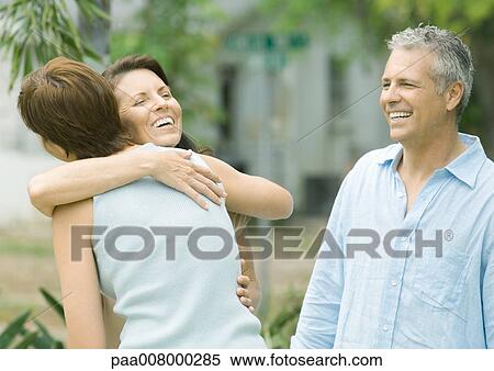Stock Photography - Two woman hugging while man watches. Fotosearch