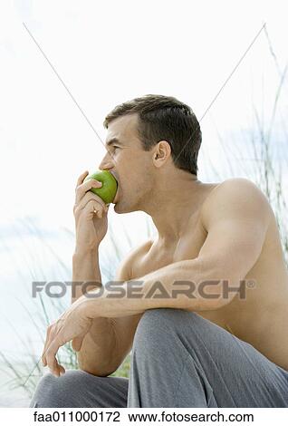 Stock Image - Barechested man eating apple, side view. Fotosearch