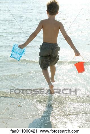 Stock Image - Boy running into surf with buckets, rear view. Fotosearch