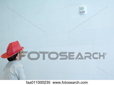 Stock Photography - Boy wearing fireman's hat, looking up at smoke alarm on wall. Fotosearch