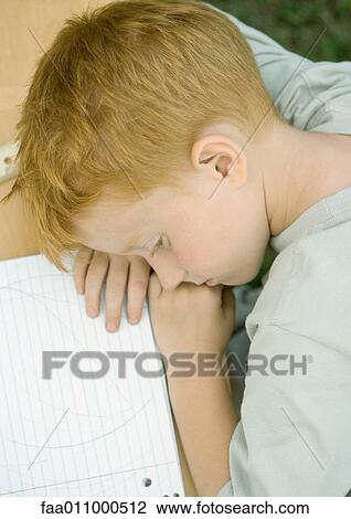 Stock Image - Boy with head down on table next to notebook, high angle view. Fotosearch