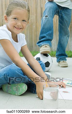 Stock Photo - Girl sitting on driveway, painting, boy with foot on soccerball in background. Fotosearch
