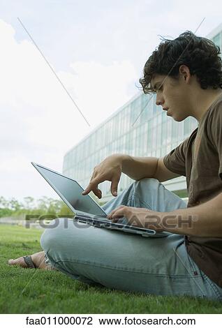 Stock Image - Male student using laptop computer in grass on campus. Fotosearch