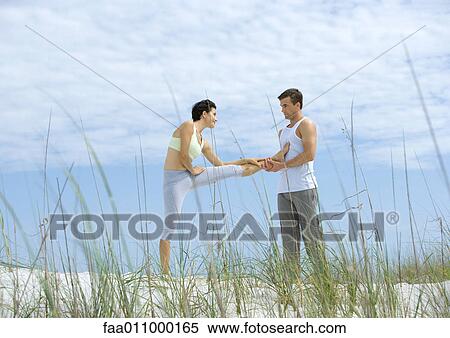 Stock Photography - Man helping woman stretch leg on beach. Fotosearch
