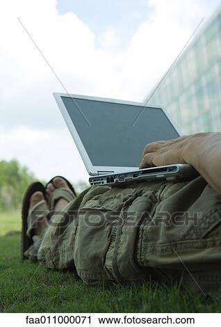 Stock Image - Man lounging in grass, using laptop. Fotosearch