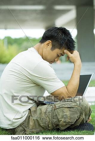 Stock Photograph - Man sitting in grass using laptop, holding head. Fotosearch
