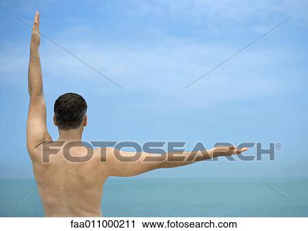 Stock Image - Man with arms making angle, sea in background, rear view. Fotosearch
