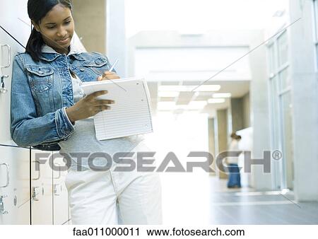 Stock Image - Teen girl standing by locker, writing in notebook. Fotosearch