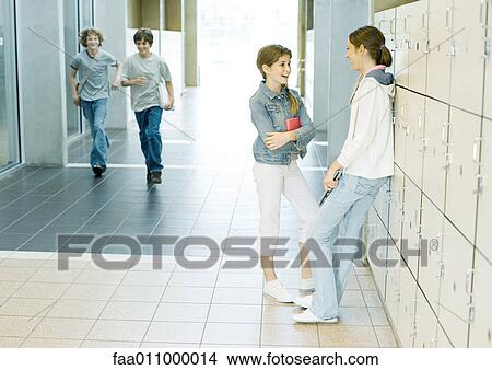 Picture - Two teen girls talking by lockers while boys run through hallway. Fotosearch