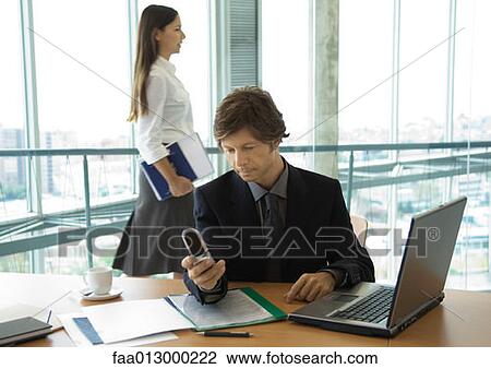 Businessman looking at cell phone, woman walking by in background View Large Photo Image Stock Image - Businessman looking at cell phone, woman walking by in background. Fotosearch