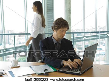 Stock Image - Businessman using laptop, woman walking by in background. Fotosearch