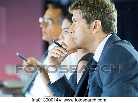 Businesspeople sitting holding pens, looking attentively out of frame View Large Photo Image Picture - Businesspeople sitting holding pens, looking attentively out of frame. Fotosearch