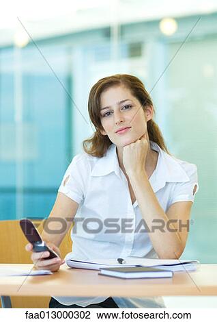 Female office worker sitting at desk, holding cell phone View Large Photo Image Stock Image - Female office worker sitting at desk, holding cell phone. Fotosearch