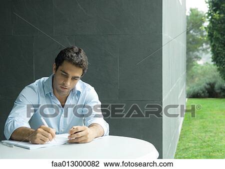 Stock Image - Man sitting at table outdoors, writing. Fotosearch