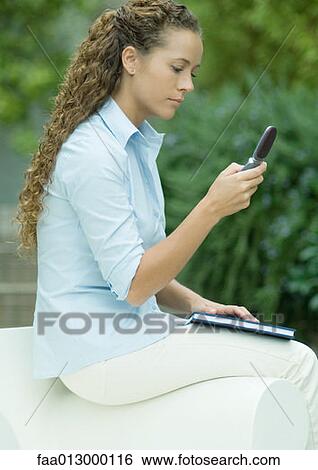 Stock Photograph - Young woman sitting on arm of chair, checking cell phone messages. Fotosearch