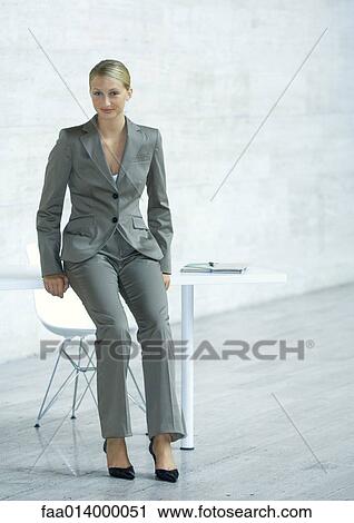 Stock Image - Businesswoman leaning against edge of table, full length portrait. Fotosearch