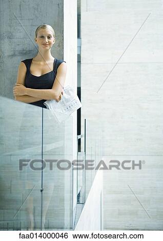 Stock Photograph - Businesswoman standing next to glass guard rail, holding newspaper, portrait. Fotosearch
