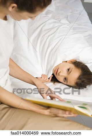 Stock Photo - Girl lying in bed, mother reading book to her. Fotosearch