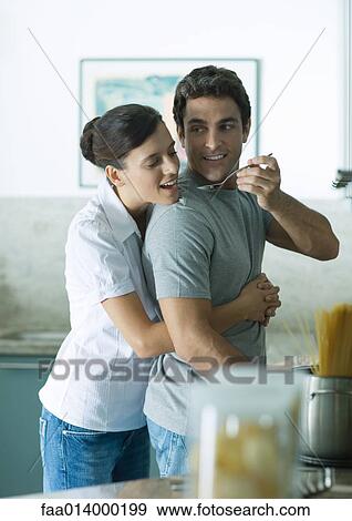 Stock Photo - In kitchen, woman holding man around waist as man reaches back with spoon for woman to taste. Fotosearch