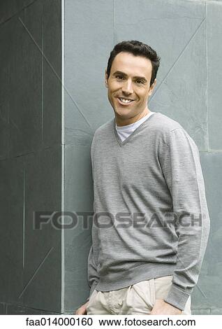 Stock Image - Man leaning against wall, smiling at camera, waist up, portrait. Fotosearch