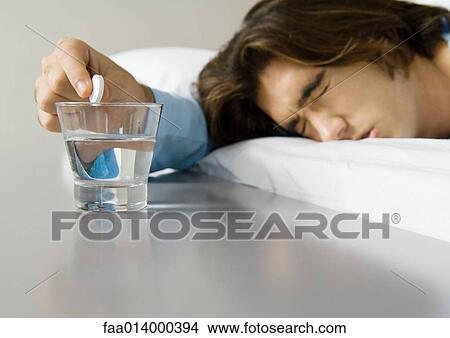 Man lying in bed, squinting eyes shut and holding effervescent medicine over glass of water View Large Photo Image Picture - Man lying in bed, squinting eyes shut and holding effervescent medicine over glass of water. Fotosearch