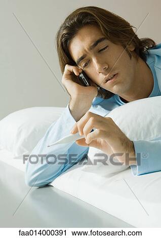 Stock Image - Man lying in bed, using cell phone and holding up thermometer. Fotosearch