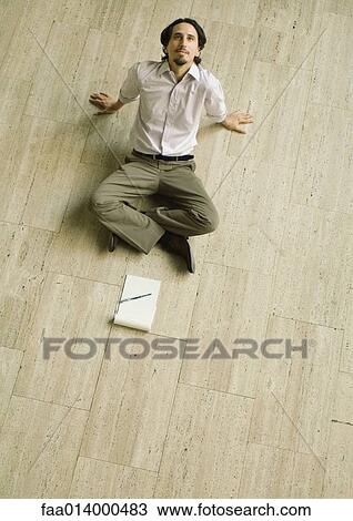 Stock Image - Man sitting on floor with pad of paper in front of him, leaning back, high angle view. Fotosearch
