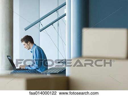 Stock Image - Man sitting on stairs using laptop, cardboard boxes in foreground. Fotosearch