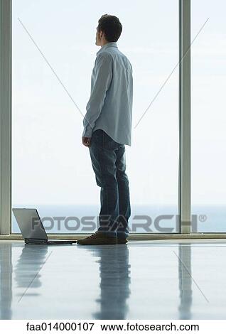 Stock Photo - Man standing, looking out bay window, laptop on floor next to him. Fotosearch