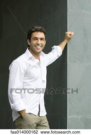Stock Image - Man standing with hand on corner of wall, smiling at camera, portrait. Fotosearch