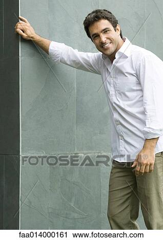 Stock Image - Man standing with hand on wall, smiling at camera, portrait. Fotosearch
