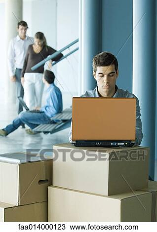 Stock Image - Man using laptop on stack of cardboard boxes, colleagues talking in background. Fotosearch