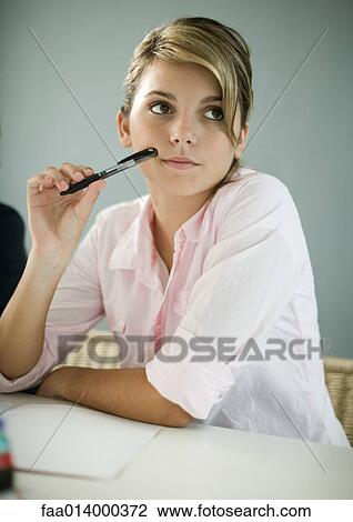 Stock Image - Teenage girl doing homework, sitting, holding pen to face. Fotosearch