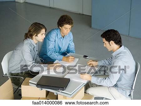 Three male colleagues sitting at table talking about documents View Large Photo Image Stock Photograph - Three male colleagues sitting at table talking about documents. Fotosearch