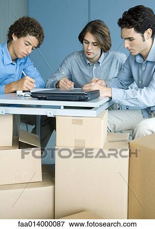 Three male colleagues working at table, cardboard boxes in foreground View Large Photo Image Stock Photo - Three male colleagues working at table, cardboard boxes in foreground. Fotosearch