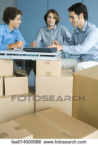 Three male colleagues working at table, cardboard boxes in foreground View Large Photo Image Stock Photo - Three male colleagues working at table, cardboard boxes in foreground. Fotosearch