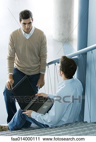 Stock Image - Two male colleagues talking, one sitting on stairs with laptop. Fotosearch