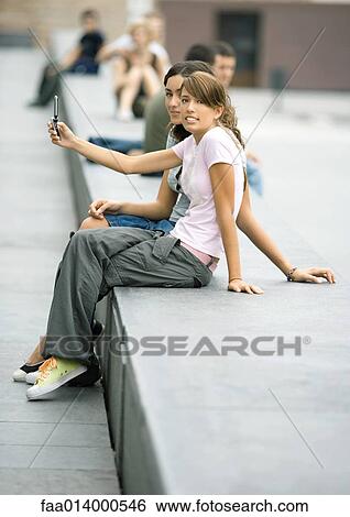 Two teenage girls sitting on bench in urban setting, taking photo with cell phone View Large Photo Image Stock Photograph - Two teenage girls sitting on bench in urban setting, taking photo with cell phone. Fotosearch
