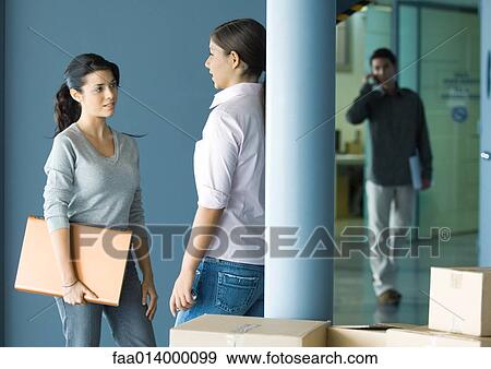 Stock Photo - Two young women standing face to face talking, man in background using cell phone. Fotosearch