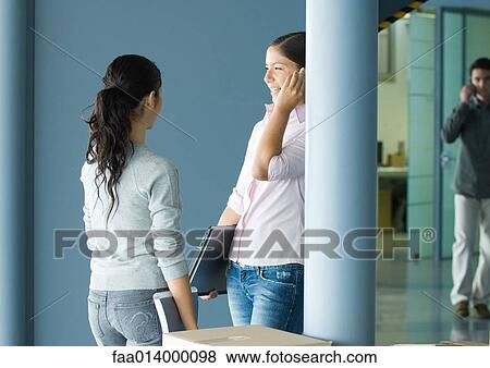 Two young women standing together near cardboard box, one using cell phone and holding laptop View Large Photo Image Stock Photo - Two young women standing together near cardboard box, one using cell phone and holding laptop. Fotosearch