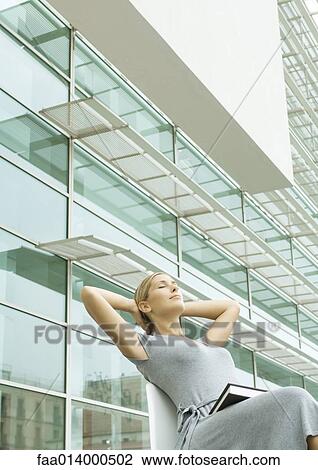 Woman sitting in chair with hands behind head, book on lap, office building in background View Large Photo Image Stock Image - Woman sitting in chair with hands behind head, book on lap, office building in background. Fotosearch
