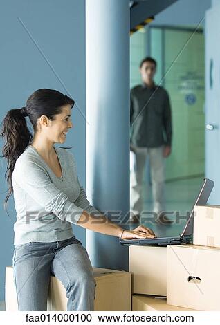 Woman sitting on cardboard box, using laptop, man standing in background View Large Photo Image Stock Image - Woman sitting on cardboard box, using laptop, man standing in background. Fotosearch