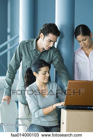 Stock Image - Woman sitting, using laptop on cardboard box, two colleagues looking over her shoulder. Fotosearch