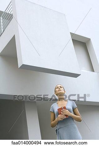 Picture - Woman standing in front of building, holding closed book, eyes shut, low angle view. Fotosearch