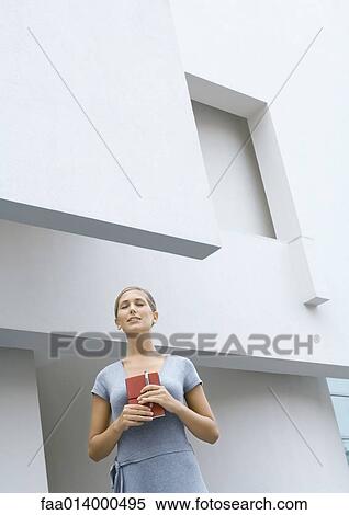 Woman standing in front of building, holding closed book, eyes shut, low angle view View Large Photo Image Stock Photography - Woman standing in front of building, holding closed book, eyes shut, low angle view. Fotosearch